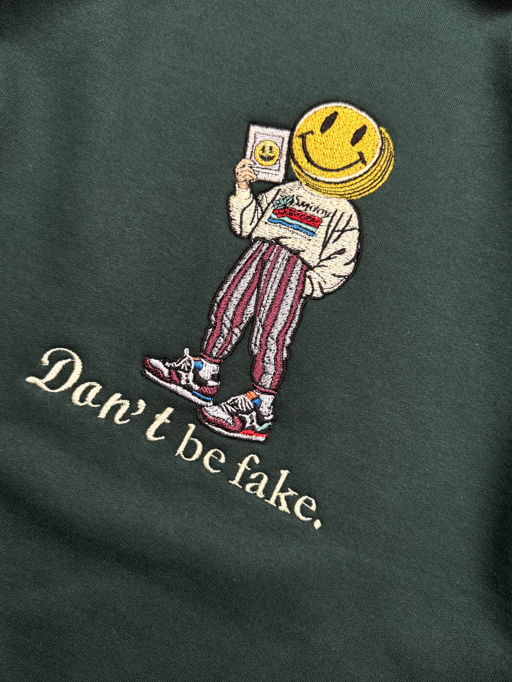 Embroidered design on a dark green fabric with a character wearing a smiley face helmet and text 'Don't be fake'.