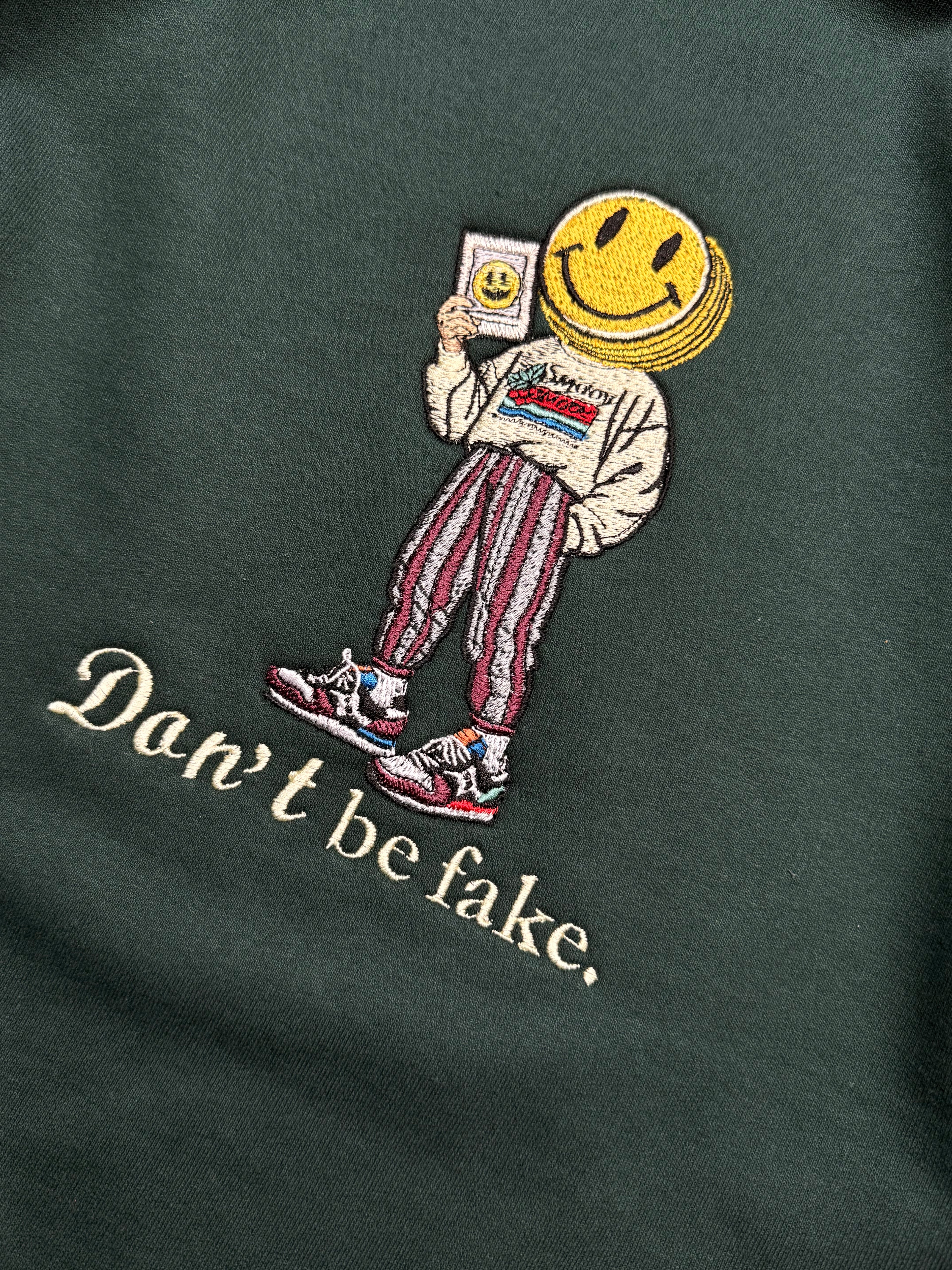 Embroidered design on a dark green fabric with a character wearing a smiley face helmet and text 'Don't be fake'.