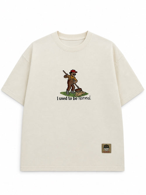 Cream embroidered tee with a cartoon man sweeping grass and the phrase "I used to be Normal"